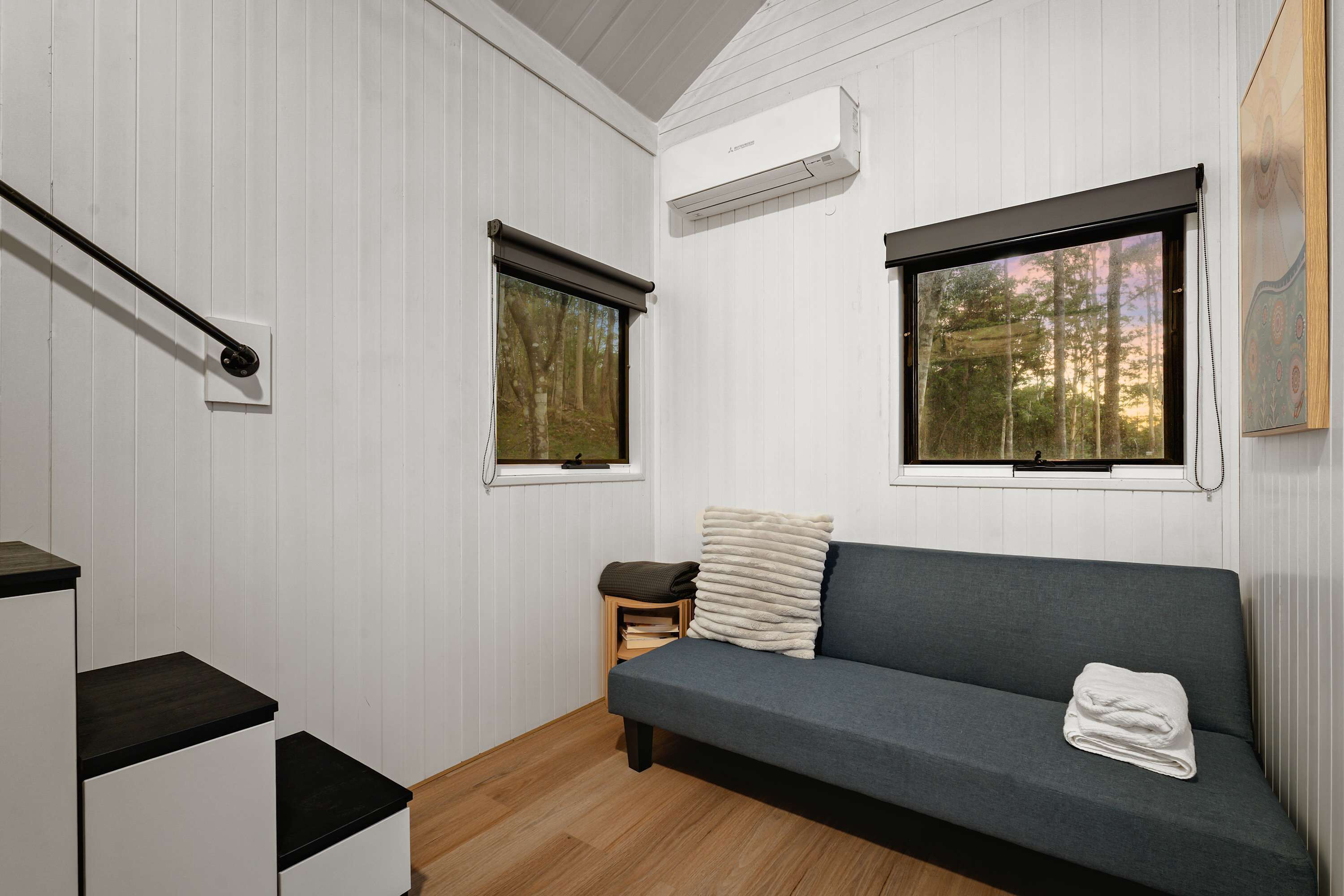 Ein modernes, kompaktes Zimmer mit einem grauen Sofa, einem kleinen Holzschemel und einem Fenster mit Blick auf Bäume.