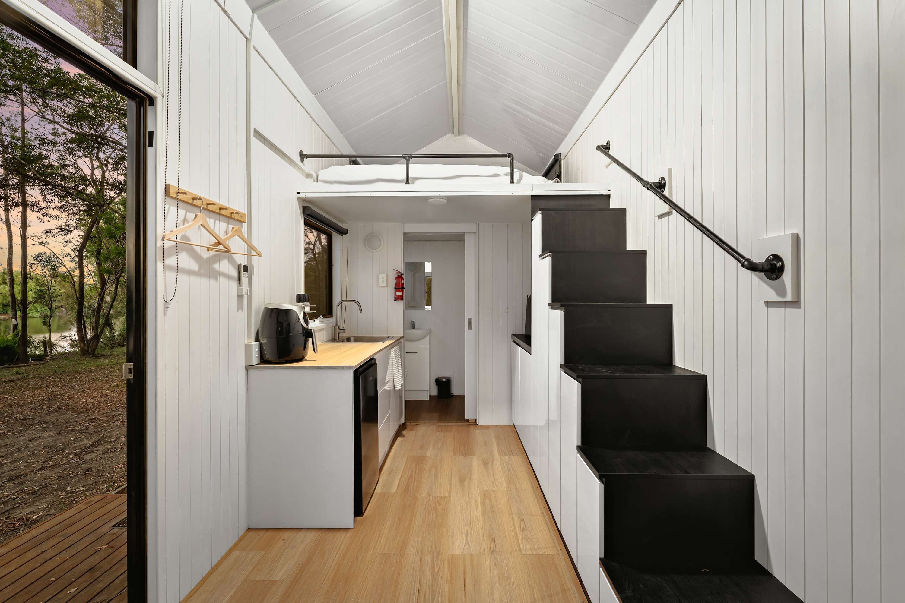 Une cuisine compacte avec un plan de travail en bois, un escalier noir et une vue sur une forêt, visible à travers une grande fenêtre.
