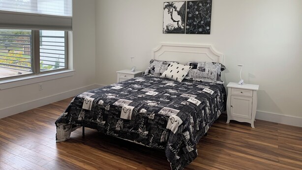 Une chambre à coucher comprenant un lit, deux tables de chevet, un ventilateur de plafond, une fenêtre avec des stores et des œuvres d’art accrochées au mur.