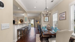 A dining area with a wooden table, chairs, and a chandelier. There is a kitchen area with a bar and stools visible in the background. - Pond-Side Retreat | Pool | Bikes | Beach Chairs & Wagon | Patio w/ Grill | Clean & Bright End Unit (St. Simons Island)