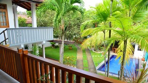 A balcony with a wooden railing overlooking a swimming pool, surrounded by lush greenery and palm trees. - Surf Villa (Hikkaduwa)