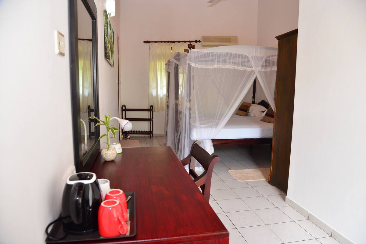 A hotel room with a wooden dining table, a bed with a canopy, a chair, and a small plant.