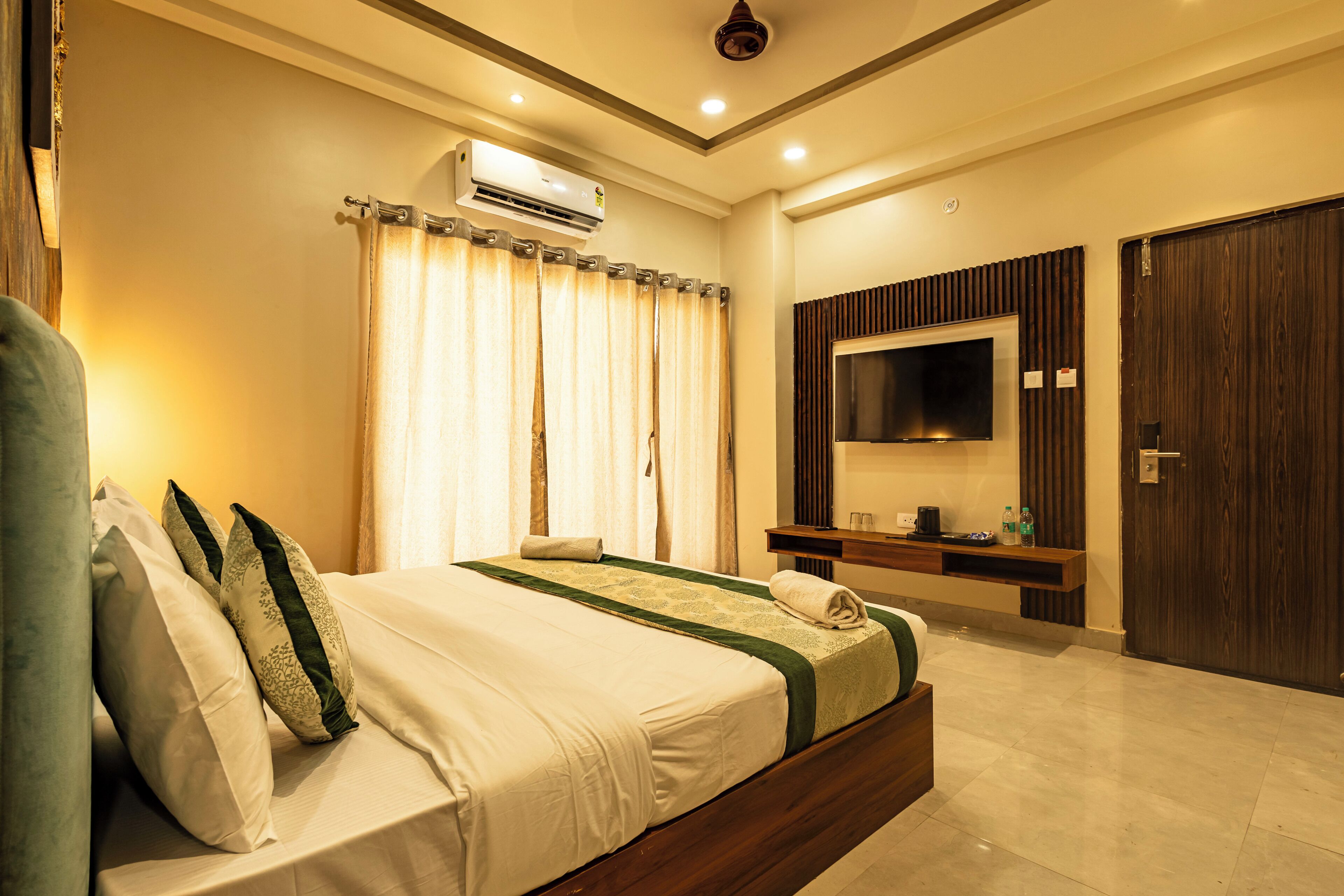 Quarto de hotel moderno com uma cama grande, ar condicionado, televisão de tela plana, guarda-roupa de madeira e uma mesa pequena com uma garrafa e copos.