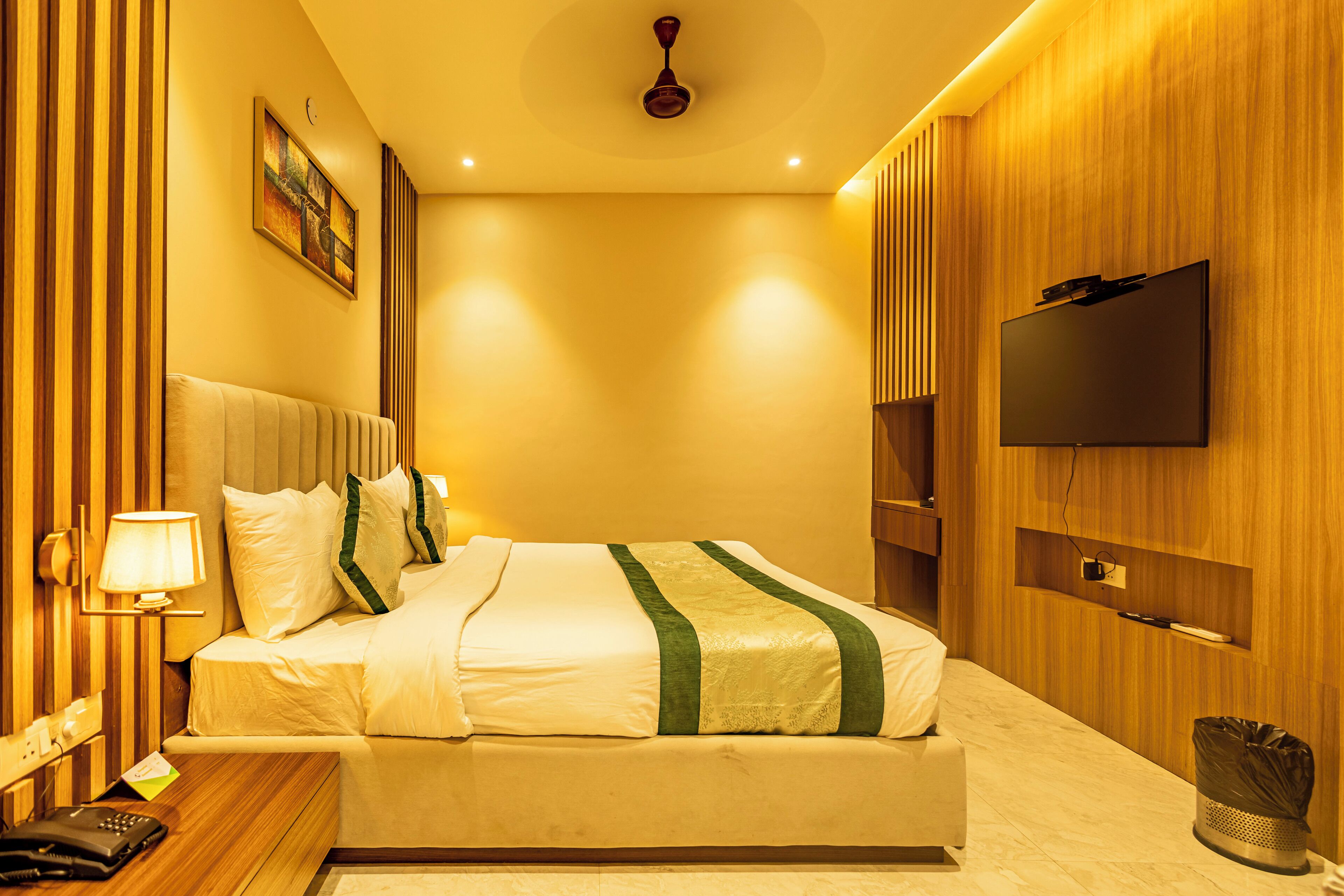 A hotel room with a bed, a TV mounted on the wall, a bedside lamp, and a trash bin.