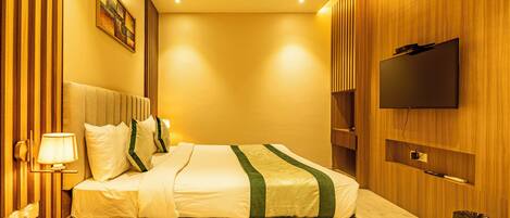 A hotel room with a bed, a TV mounted on the wall, a bedside lamp, and a trash bin.