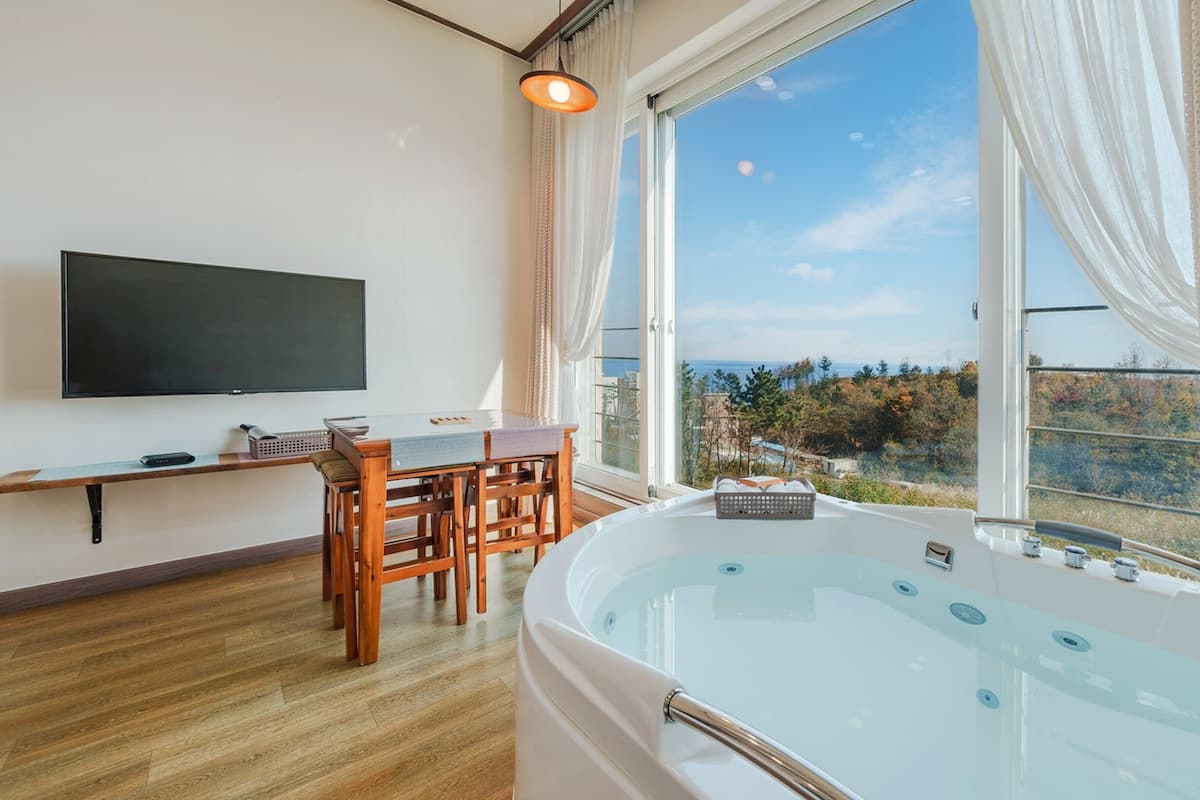 Een kamer met een groot raam, een houten tafel, stoelen, een flatscreen televisie en een whirlpool.