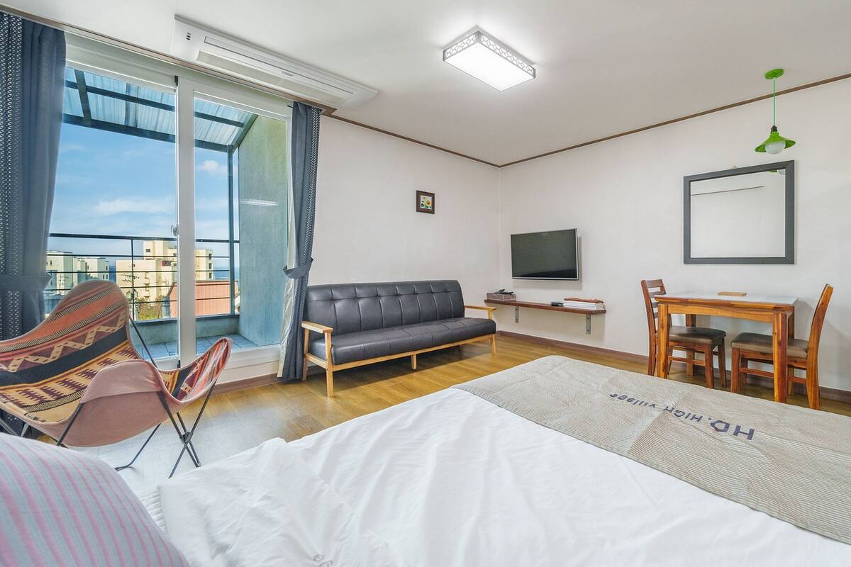 Een hotelkamer met een bank, een eettafel met stoelen, een televisie, een koelkast en een groot raam met uitzicht op gebouwen.