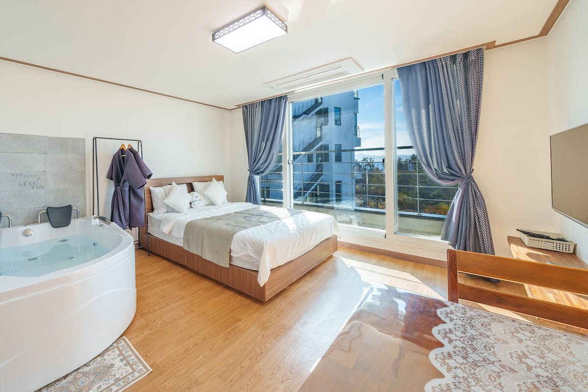 Een moderne slaapkamer met een groot bed, een aparte badkamer met jacuzzi, en uitzicht op de stad door grote ramen.