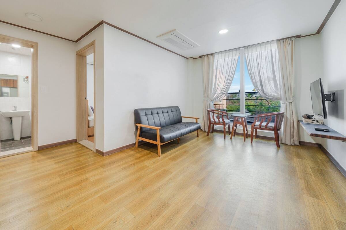 Een ruime woonkamer met een bank, eettafel en stoelen, een aan de muur bevestigde televisie, en uitzicht door grote ramen.