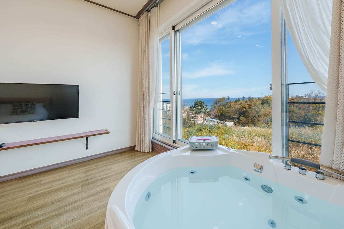 Een moderne badkamer met een ruime badkuip, een aan de muur bevestigde televisie en een raam met uitzicht op de zee en bomen.