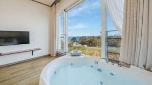 A modern bathroom with a large bathtub, a TV mounted on the wall, and a window offering a scenic view of the sea and trees. - Gangreung top spa Pension (Gangneung)