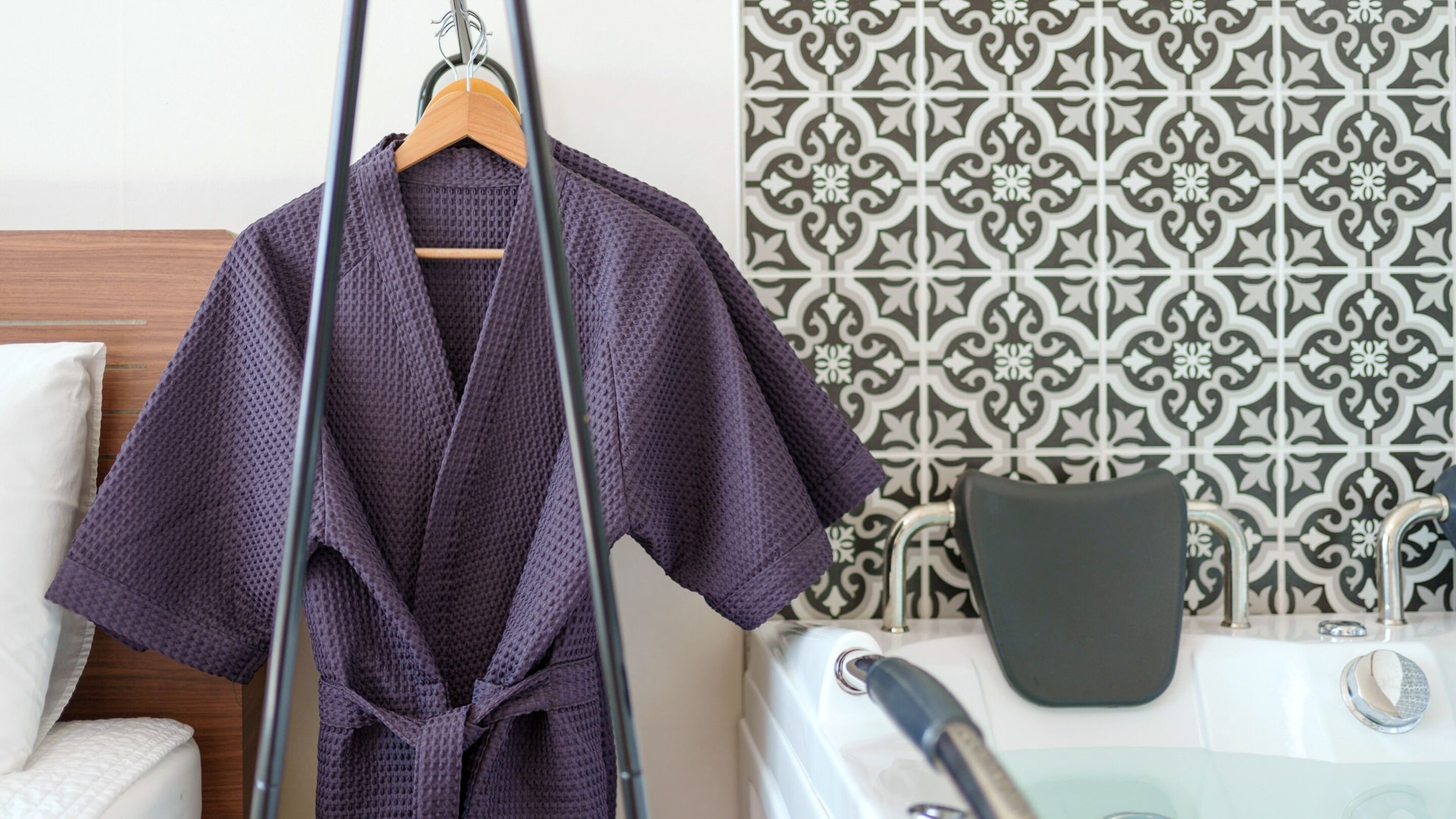 Une robe violette est suspendue à un portant, un oreiller blanc est posé sur un lit, et un mur à motifs se trouve dans une pièce qui contient également une baignoire.