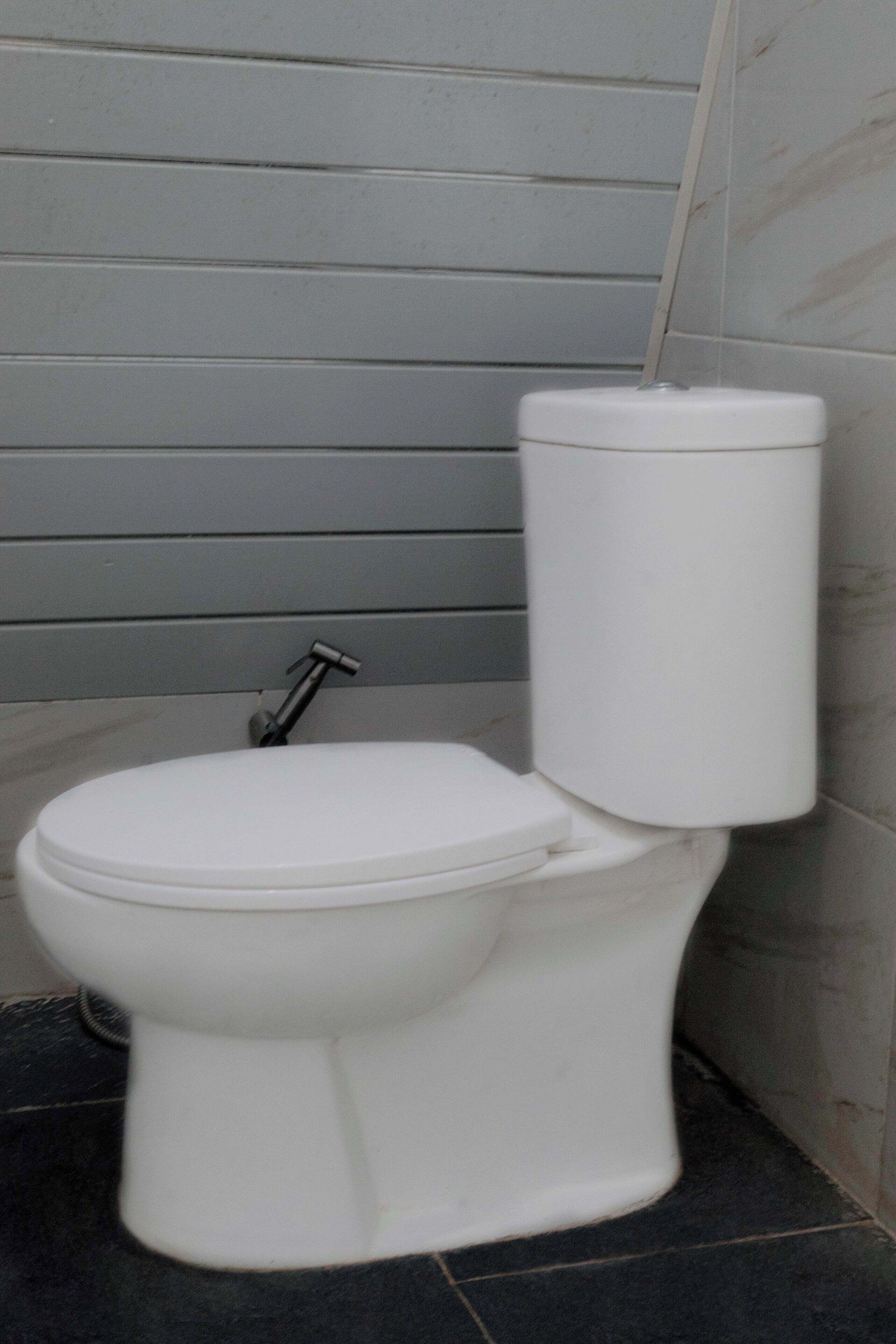 A white toilet with a tank and a flush handle in a bathroom with grey tiled walls and black floor tiles.