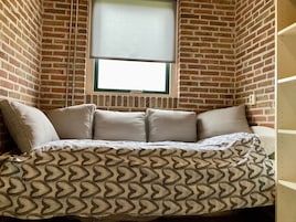 A corner room with a brick wall, a window with a blind, and a bed with patterned bedding.
