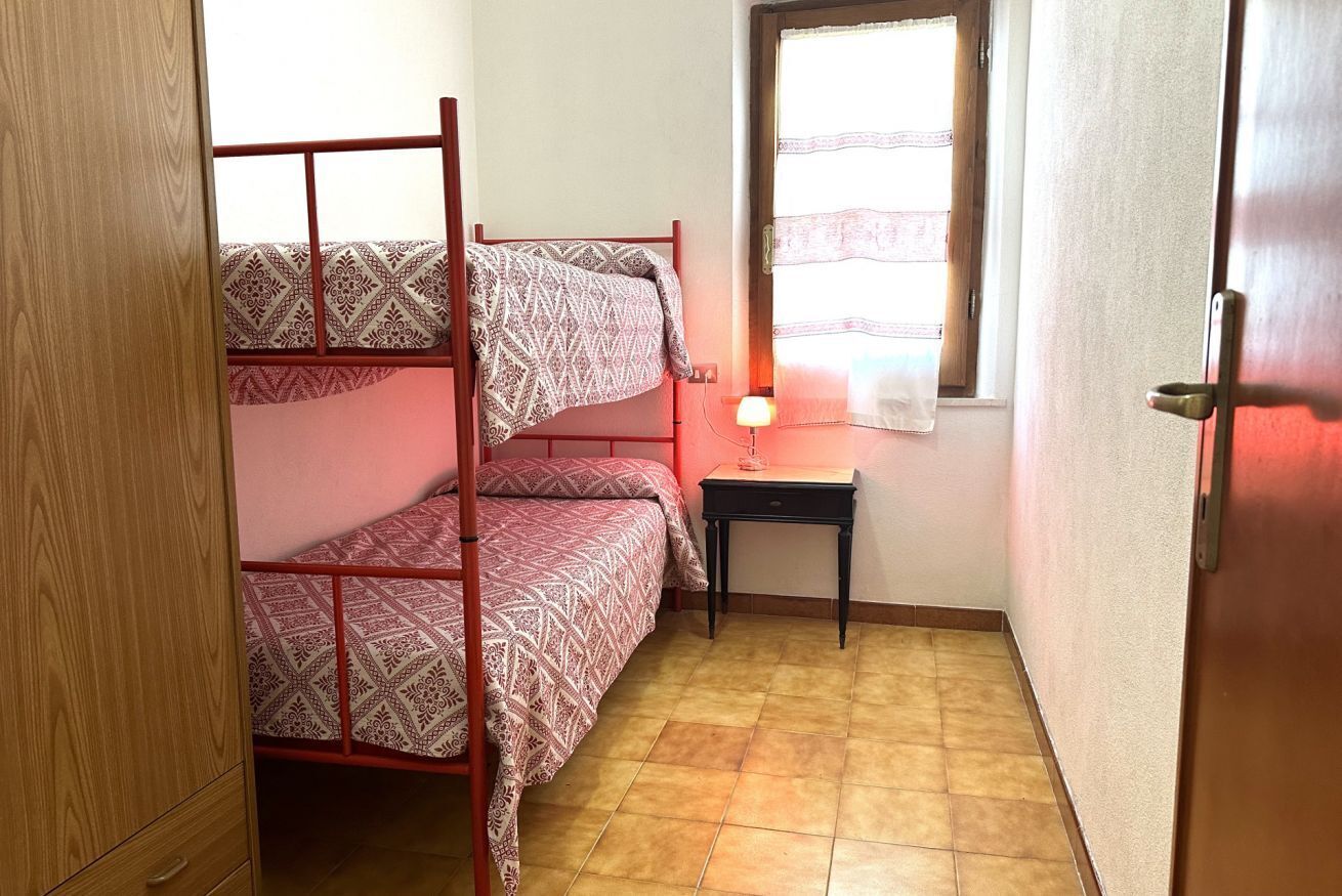Een studentenkamer met stapelbedden, een klein tafeltje met een lamp, een raam met gordijnen en een houten garderobe.