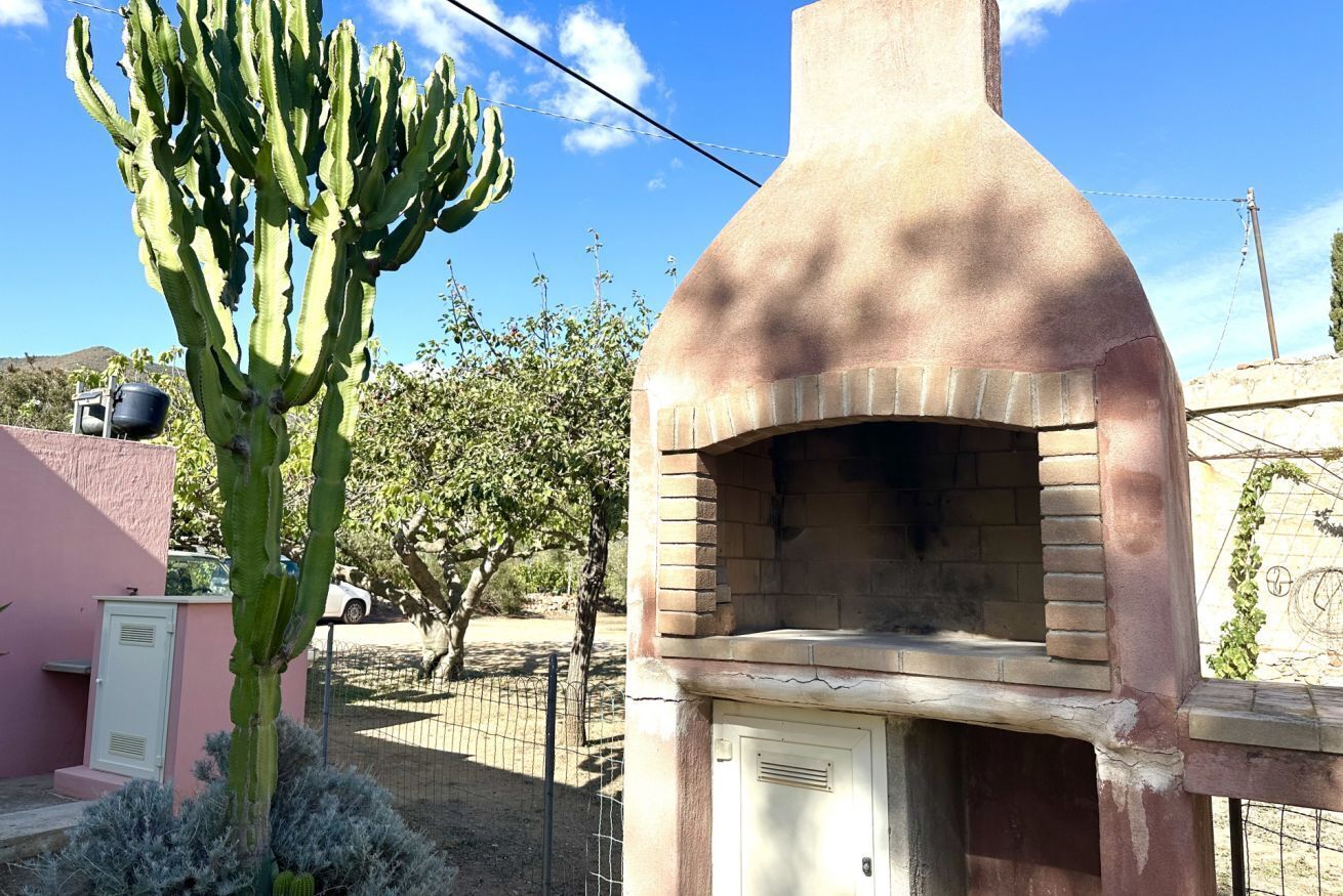 Estructura de ladrillo con un horno y una puerta, rodeada de una valla y follaje.