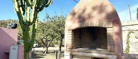 Estructura de ladrillo con un horno y una puerta, rodeada de una valla y follaje.