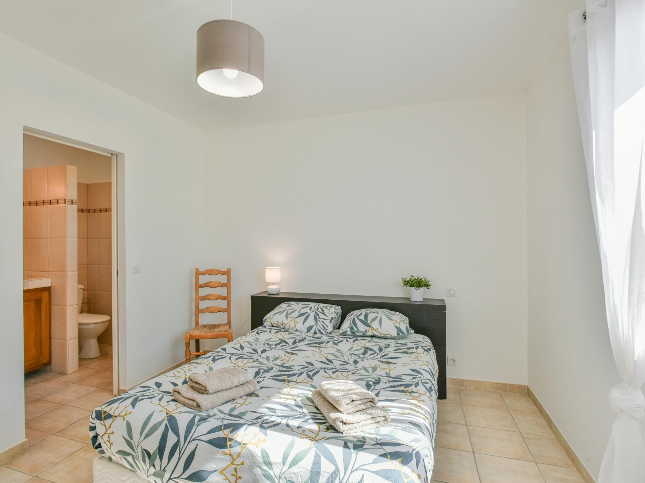 A bedroom with a bed, a bedside table with a lamp, a chair, a potted plant, and a bathroom visible through an open door.
