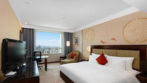 Habitación de hotel con una cama grande, un escritorio con televisor, una silla y vistas a la ciudad a través de un ventanal.
