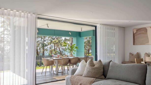 A modern living room with a grey sofa, white curtains, and a view of an outdoor dining area. - The Bowen Huskisson (Huskisson)