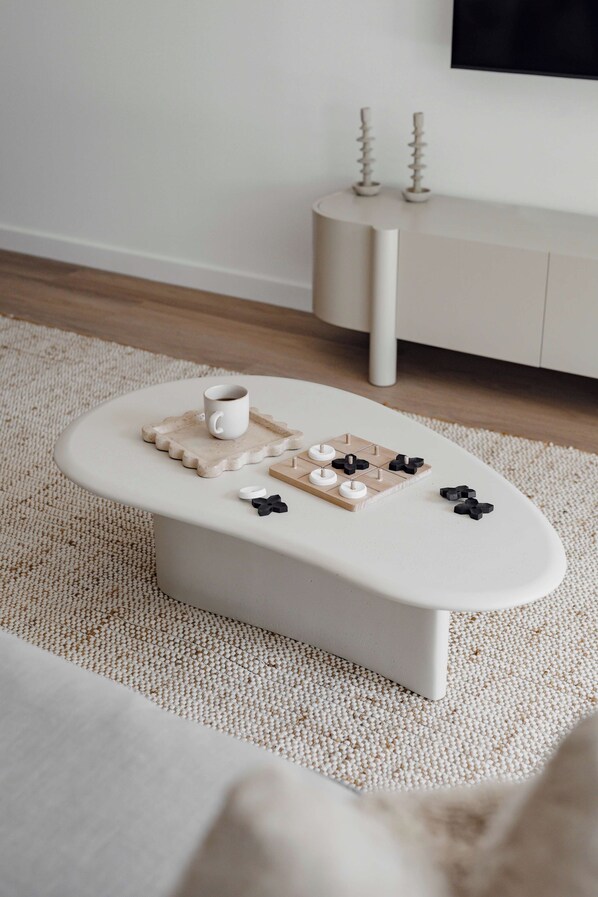 A white, oval-shaped coffee table with a coaster and a cup on it, a board game, and two candle holders on a beige rug. - The Bowen Huskisson (Huskisson)