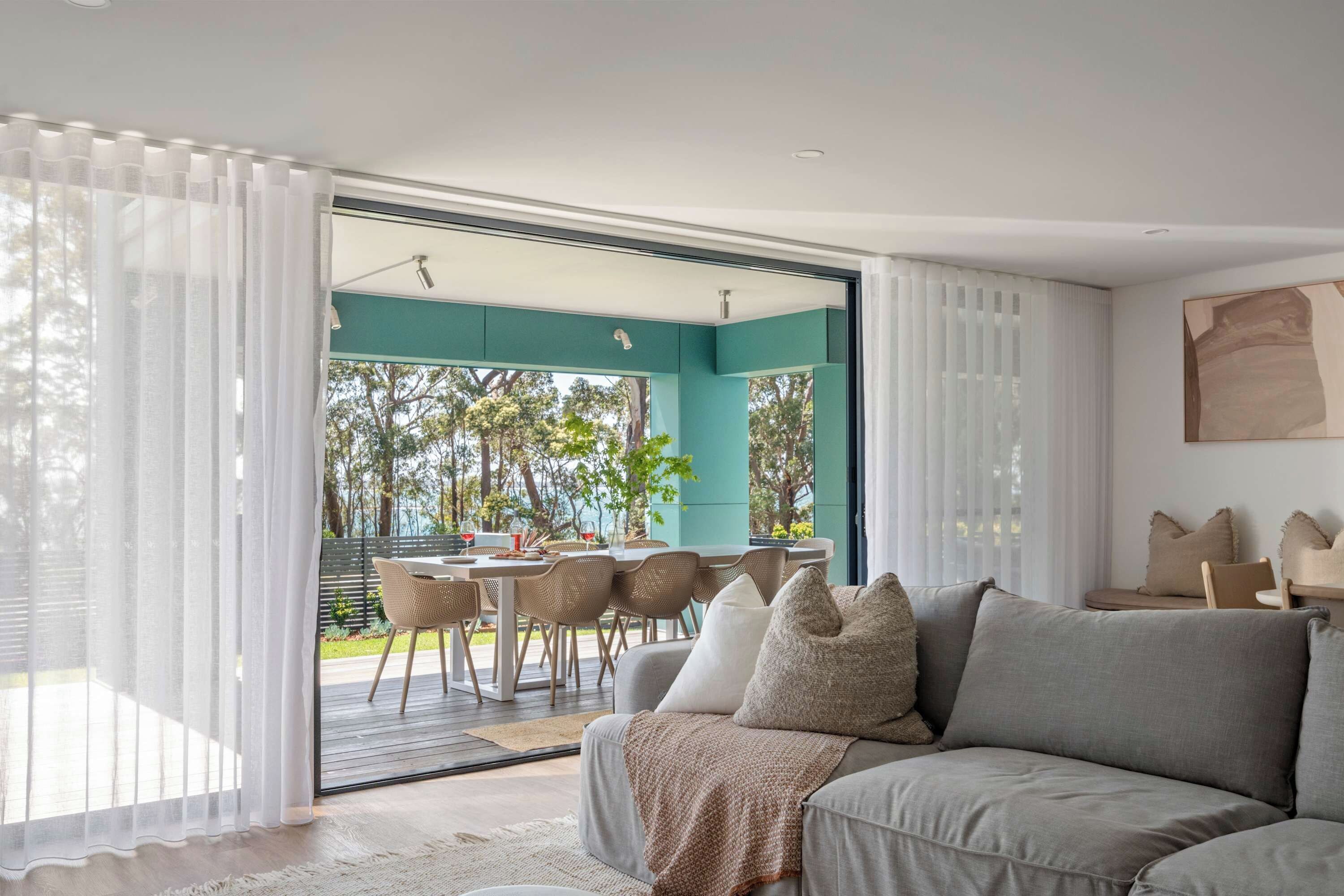 A modern living room with a grey sofa, white curtains, and a view of an outdoor dining area.