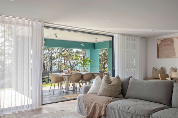 A modern living room with a grey sofa, white curtains, and a view of an outdoor dining area.