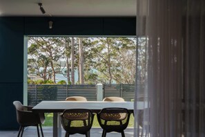 A dining area with a table and chairs, a view of trees and a body of water through a glass partition. - The Bowen Huskisson (Huskisson)