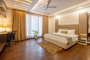 A modern hotel room with a large bed, a desk, a chair, a ceiling fan, and a large window with curtains. - Vivid Boutique Hotel, Medanta, Gurugram (Gurugram)