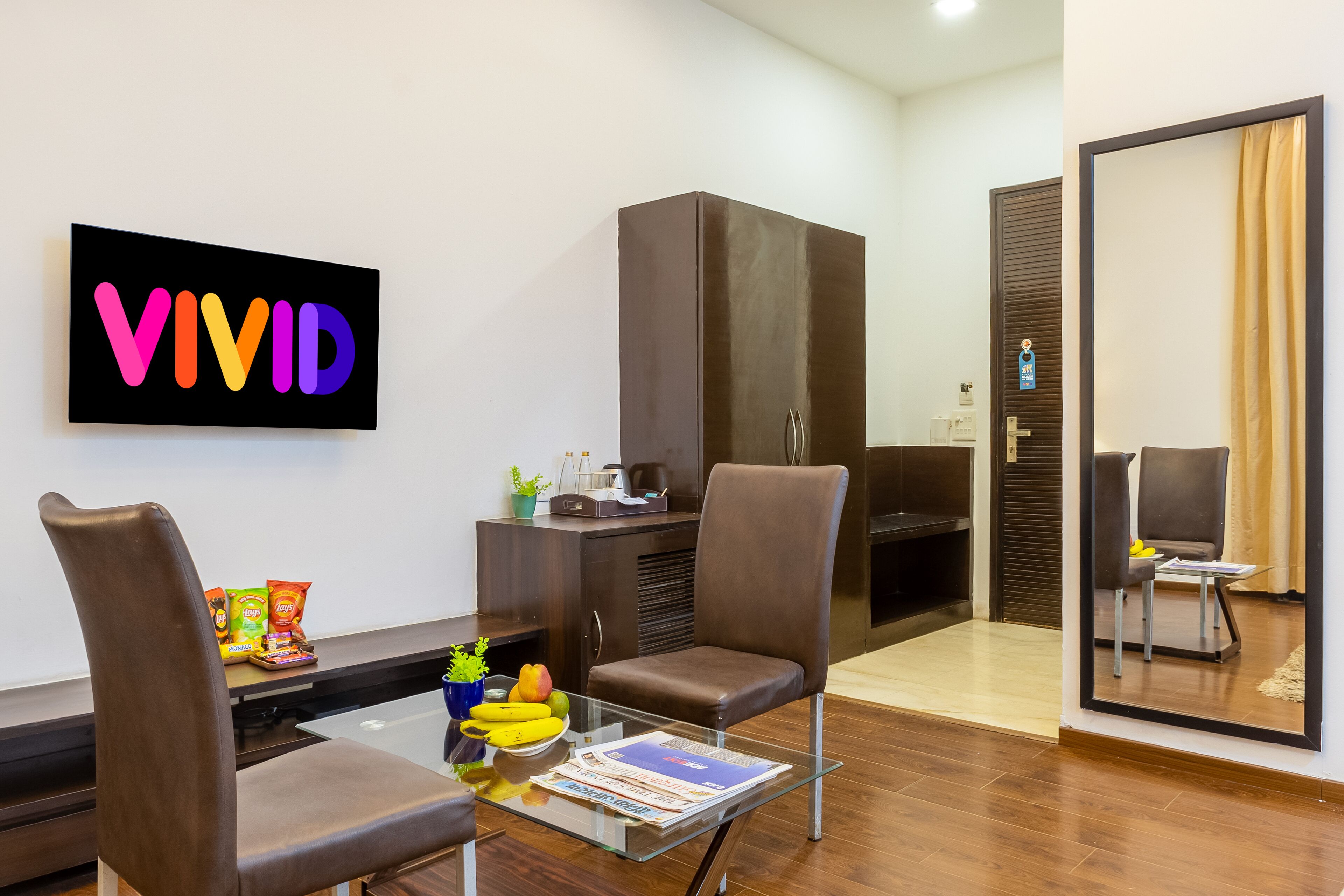 A modern hotel room with a glass table, two brown chairs, a dark wooden wardrobe, and a large mirror.