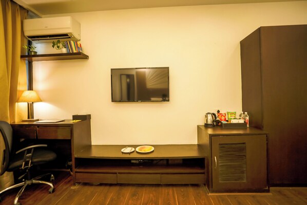 A hotel room with a TV mounted on the wall, a desk with a chair, a small kitchenette, and a wooden floor. - Vivid Boutique Hotel, Medanta, Gurugram (Gurugram)