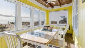 A bright yellow room with white walls and wooden ceiling beams, featuring a table with chairs, large windows, and a view of the beach. - The Ocean Rose - Classic Dog Friendly Beachfront Gem with Large Sunroom and Outdoor Deck (Oak Island)