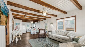 A spacious living room with a large sofa, a coffee table, and a patterned rug. There is a dining area with a table and chairs, and a kitchen in the background. - The Ocean Rose - Classic Dog Friendly Beachfront Gem with Large Sunroom and Outdoor Deck (Oak Island)