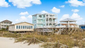 A beachfront property with multiple buildings, including a prominent two-story house with a balcony and a smaller adjacent structure. - The Ocean Rose - Classic Dog Friendly Beachfront Gem with Large Sunroom and Outdoor Deck (Oak Island)