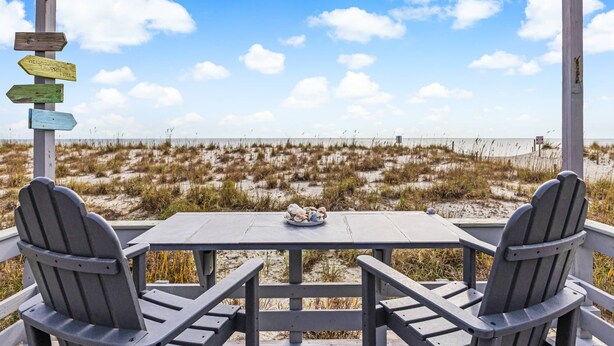 A beachside table with two chairs and a plate of seashells, overlooking a beach with dunes and a wooden signpost. - The Ocean Rose - Classic Dog Friendly Beachfront Gem with Large Sunroom and Outdoor Deck (Oak Island)