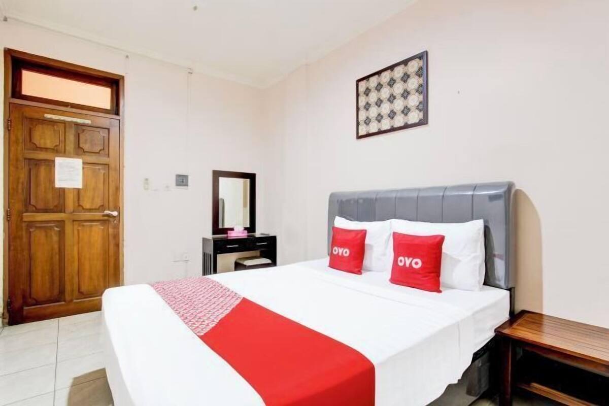 A hotel room with a bed, a wooden door, a mirror, a bedside table, and a wall-mounted picture.