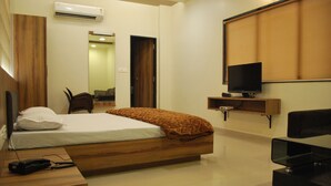 A modern bedroom with a wooden bed, a TV on a wooden stand, a desk with a phone, and a mirror on the wall. - Hotel Vijya laxmi Rooms and Banquet Hall (Surat)