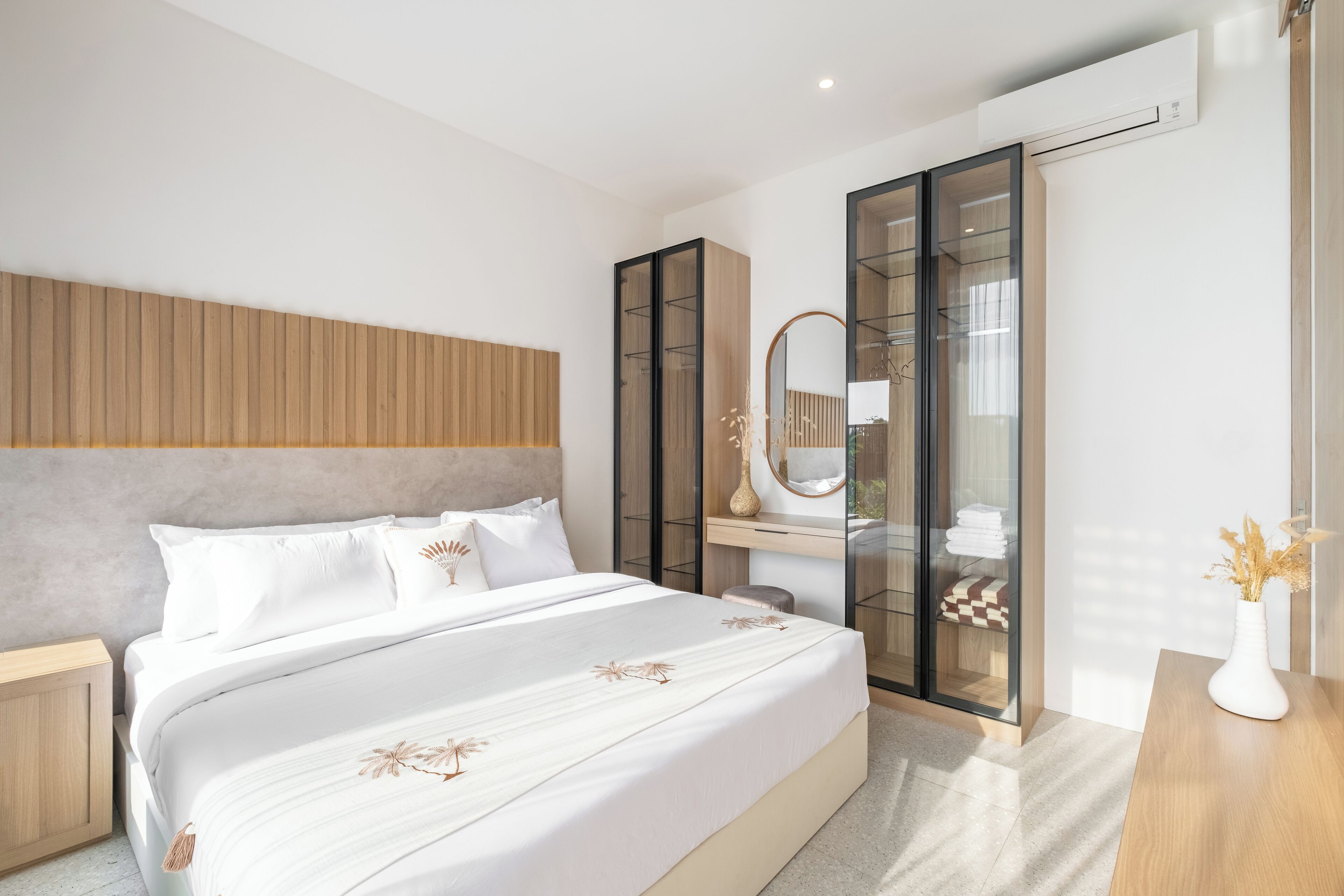 A modern bedroom with a large bed, a wooden headboard, a glass wardrobe, and a bedside table with a vase.
