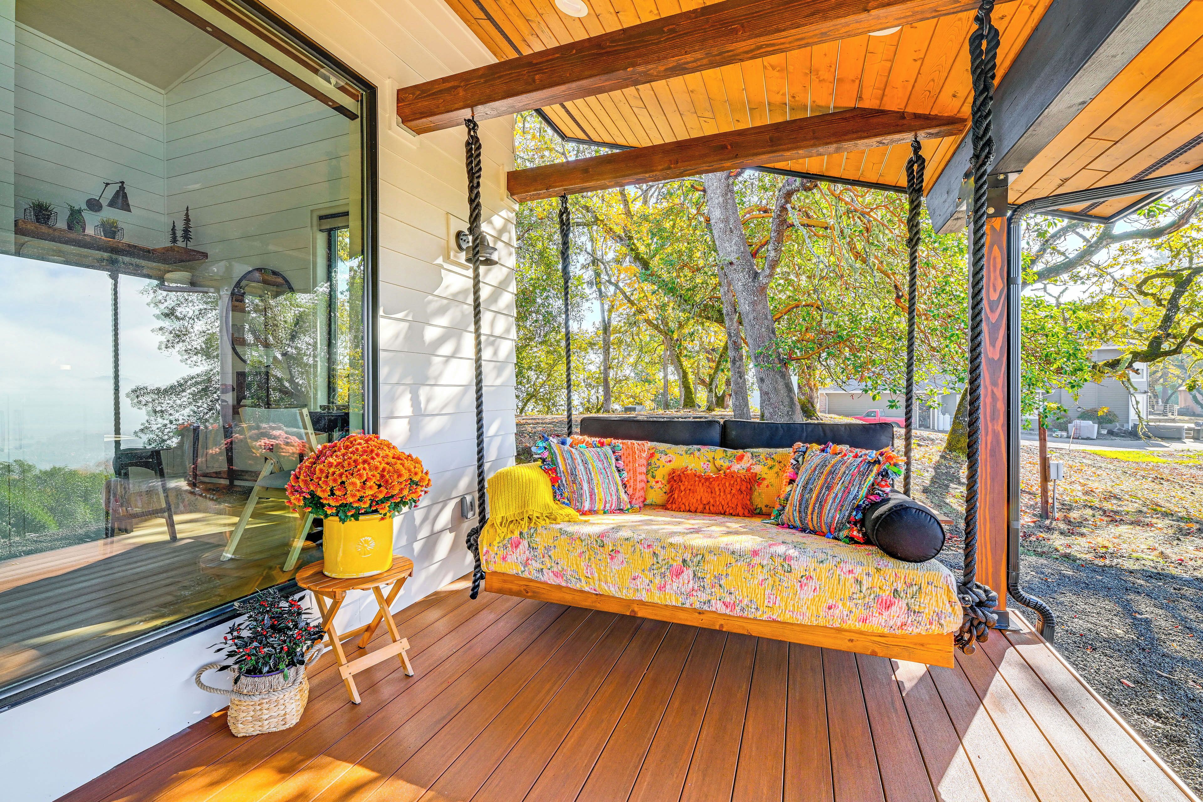 Una terraza de madera con una hamaca colgante, una maceta amarilla con flores y vistas a los árboles a través de amplias ventanas.