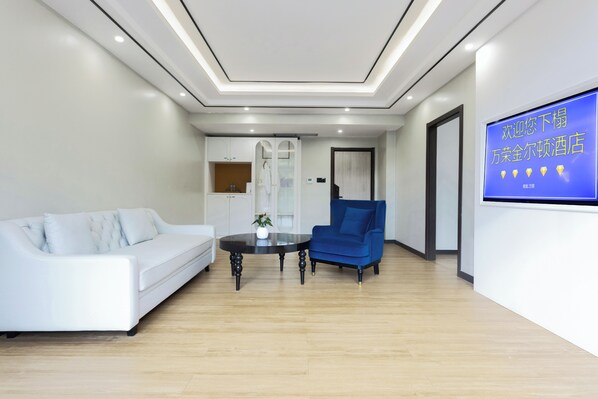 Quarto de hotel moderno com um sofá branco, uma poltrona azul, uma mesa redonda com um vaso e uma televisão exibindo texto.