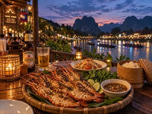 A dining table with grilled seafood, a beer bottle, and a glass of beer, set against a backdrop of a water body and mountains at dusk.