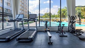 Een sportschool met loopbanden, een elliptische trainer en een roeimachine, met uitzicht op een zwembad en een gebouw met balkons.