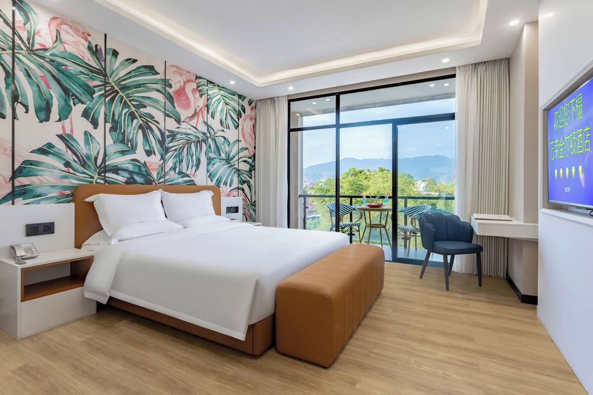 A modern hotel room with a large bed, a nightstand, a TV, and a balcony with a view of the mountains.