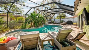 A covered outdoor swimming pool with lounge chairs, a barbecue area, and a view of a house.