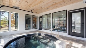 A modern indoor swimming pool with a wooden ceiling, large windows, and a stone border. - Luxury 3 Br Cabin~Indoor Pool~Hot Tub~Fire Pit (Sevierville)