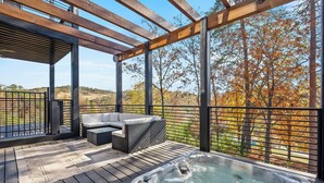 A wooden deck with a hot tub, outdoor furniture, and a view of autumn trees. - Luxury 3 Br Cabin~Indoor Pool~Hot Tub~Fire Pit (Sevierville)