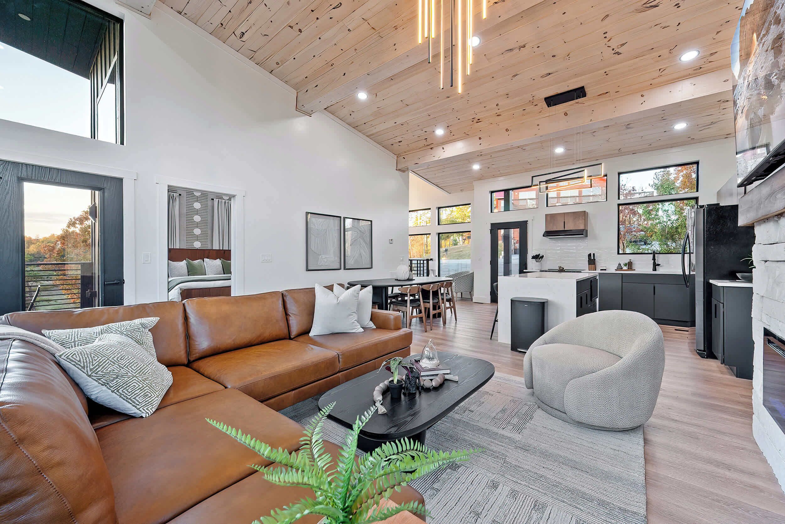 A modern living room with a brown leather sofa, a black coffee table, and a grey armchair. There is a kitchen in the background with a dining area.