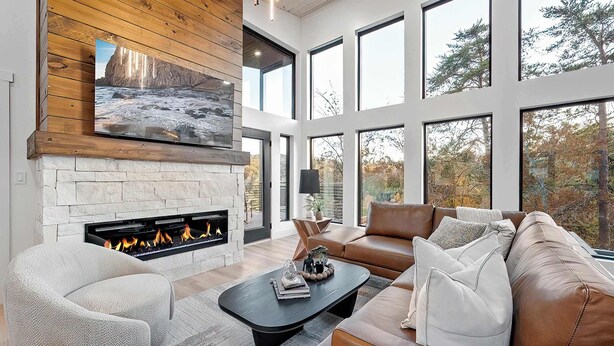 A modern living room with a stone fireplace, leather sofas, and large windows offering a view of trees. - Luxury 3 Br Cabin~Indoor Pool~Hot Tub~Fire Pit (Sevierville)
