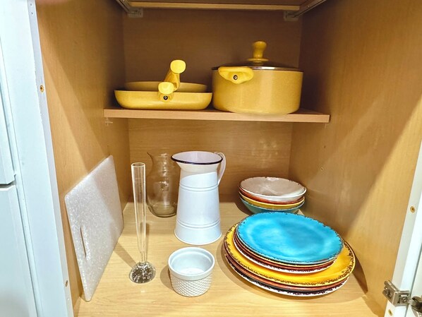 A kitchen with a wooden shelf holding a yellow pot and a yellow lid, a white pitcher, a clear glass vase, and a stack of colorful plates. - Pauper's Palace (Prescott)