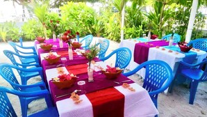 An outdoor dining area with tables set for multiple guests, surrounded by blue chairs and covered with tablecloths.